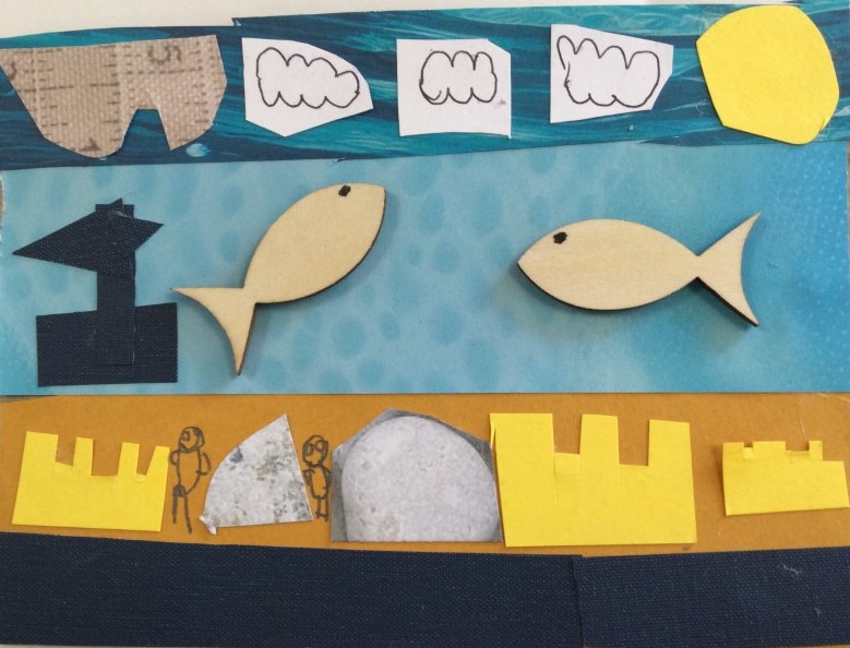 A postcard collaged with paper and wooden shapes, with a black border along the bottom, above which are cut-out shapes of sandcastles, stones and hand drawn people. Above it a strip of blue paper with two wooden fish glued and a black paper boat. The sky above is created with a strip of blue patterned paper, overlaid with white clouds and a yellow sun