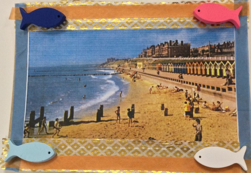 A collaged postcard, with an image of children in swimsuits playing on a sandy beach against a background of beach huts and large hotels, with a border in gold and blue with a blue wooden fish top left, a pink wooden fish top right, a light blue wooden fish bottom left and a plain wooden fish bottom right