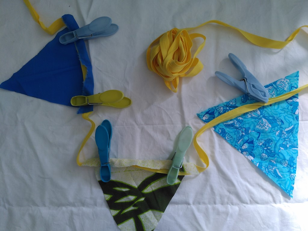 A plain blue flag is pegged to a length of yellow tape, using a blue peg and a yellow peg. A second flag of plain calico with a swirling green line on it is also pegged to the tape and a final flag, this time light blue patterned, has the tape running across it and a peg ready to fix it to the tape