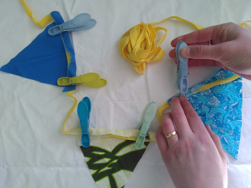 A pair of hands is shown, pegging a bunting flag onto the yellow tape. Two other flags are already in position and pegged