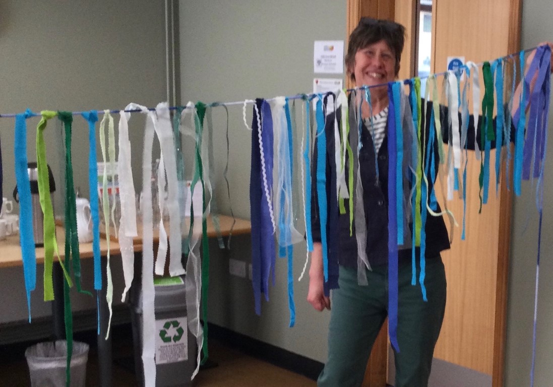 A person stands in front of a closed door, in one hand holding a string of bunting made out of lots of strips of fabrics in different shades of blue and green