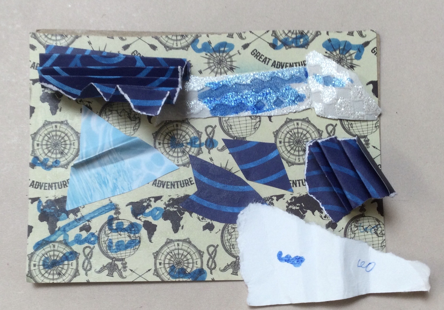 A postcard collaged with a background of paper showing compasses, globes and the words Great Adventure, overlaid with blue patterned papers, some of them folded like concertinas to represent waves