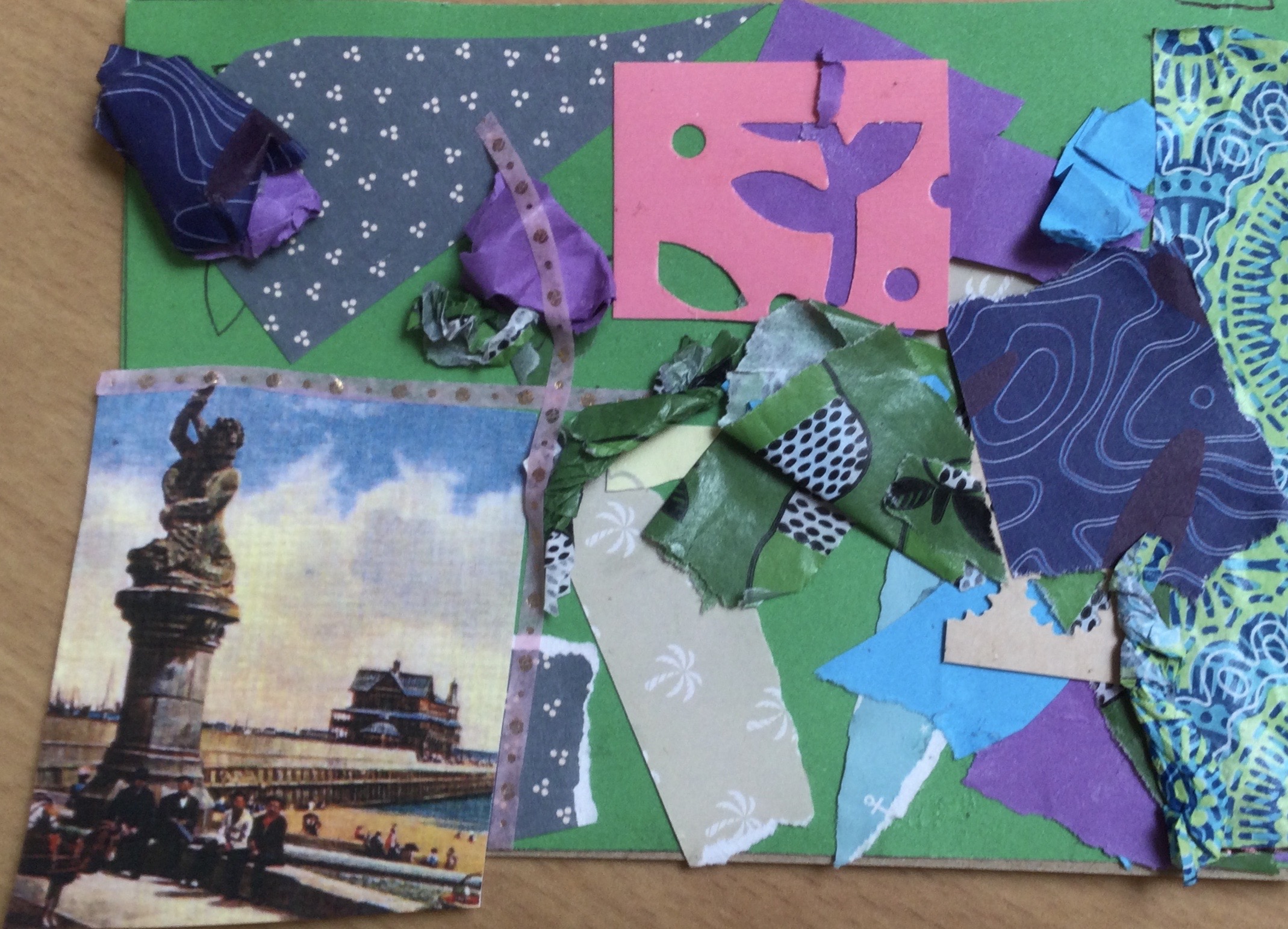 A collaged postcard made with a variety of blue and green patterned papers, thin tape and a piece of pink latticed card. A snippet from a vintage postcard showing the pier and statue of Titan is bottom right.