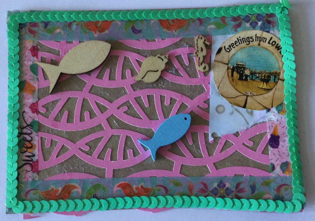 A brown postcard collaged with pink latticed paper, strips of tape and an image from an old postcard saying Greetings from Lowestoft. Small wooden fish, a shell and a seahorse are glued on top and the card has a border of green sequins