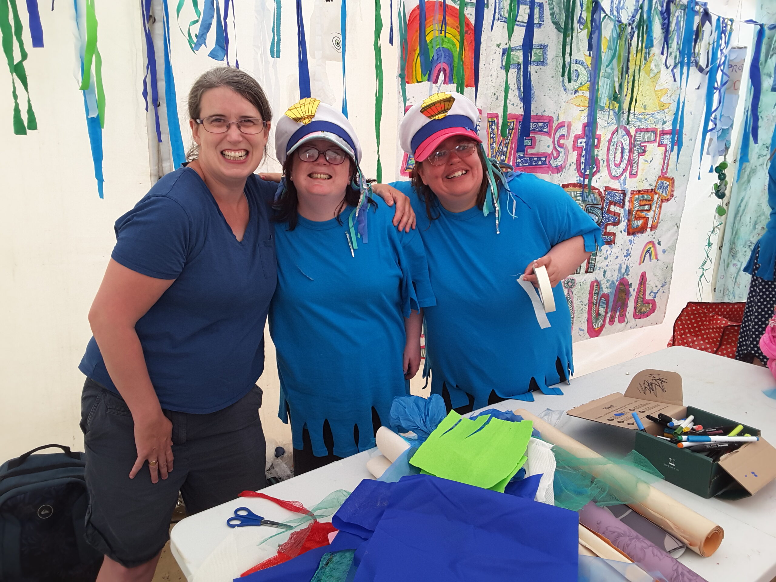 Three people, all wearing shades of blue and two wearing sailors' caps with gold crown emblems on them, stand with their arms over each others' shoulders, grinning at the camera. Behind is a festoon of bunting and a painted fabric banner and on a table in front of them are materials, papers and pens - all ready for people to make their mythical sea creatures
