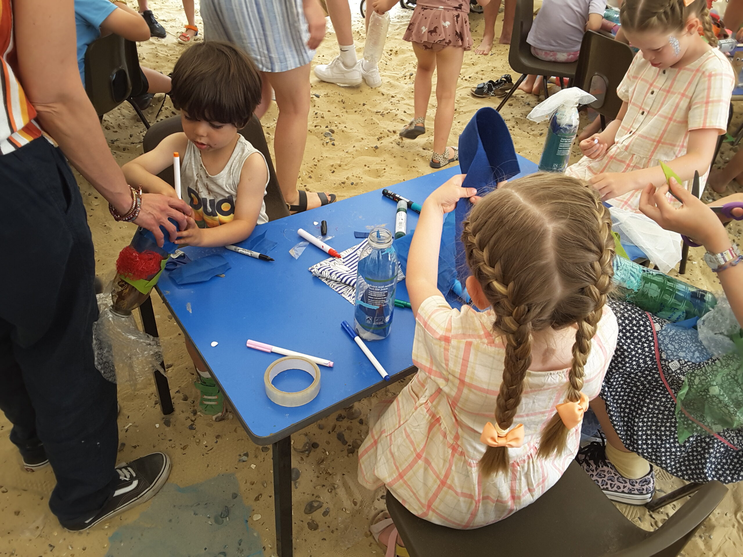 Young children seated at a small, blue-topped table, surrounded by pens, Sellotaphe and other materials, making sea creatures out of recycled materials