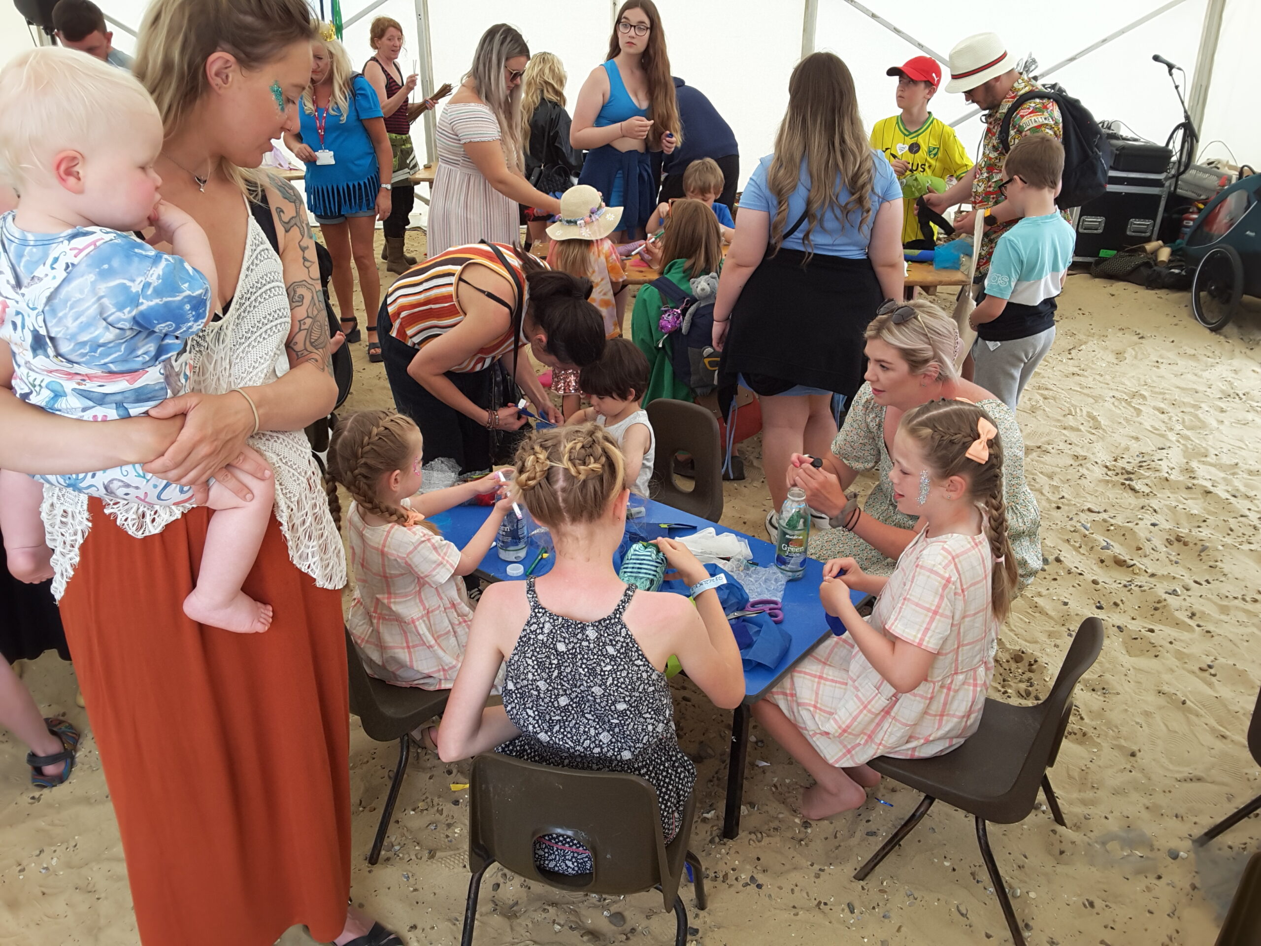 The crowded interior of a large marquee, in which families are seated and standing, making objects about of recycled plastic bottles and other materials