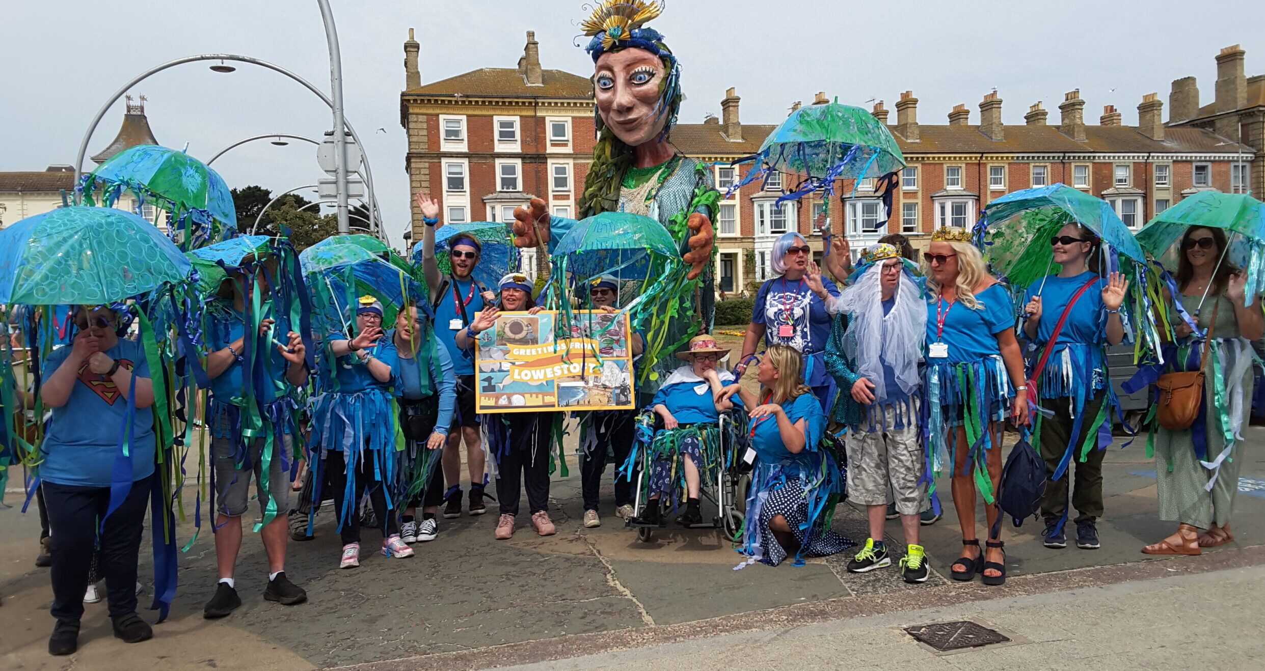 A gigantic puppet with a rising sun gold crown stands behind a line of people, dressed in bright blue T-shirts and carrying umbrellas decorated to look like sea creatures. In the centre, two participants are holding a large postcard with the message Greetings from Lowestoft.