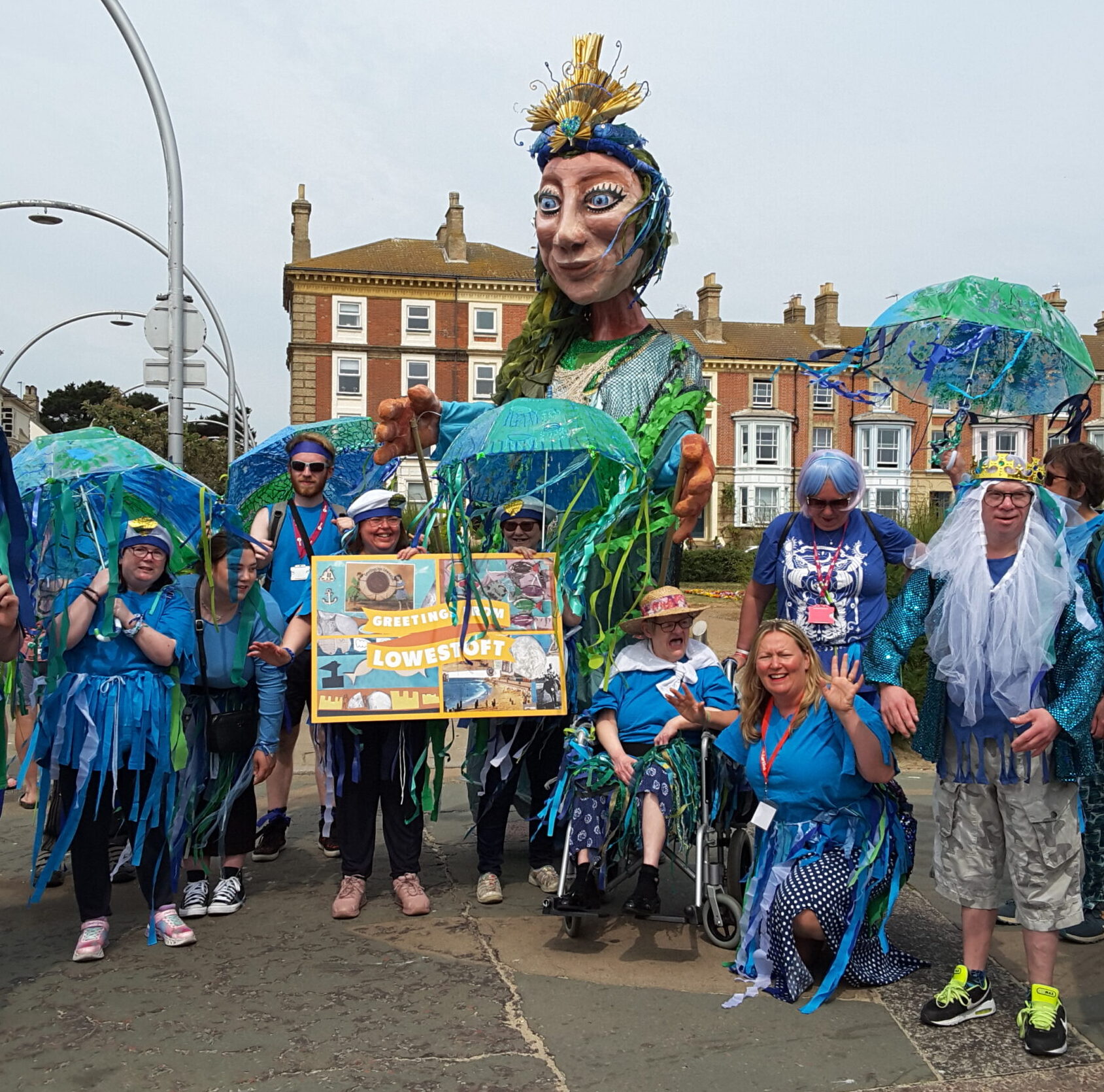 The Brave Artists, dressed in fancy dress to resemble sea creatures, greet the massive puppet, Sol, with an equally outsized postcard