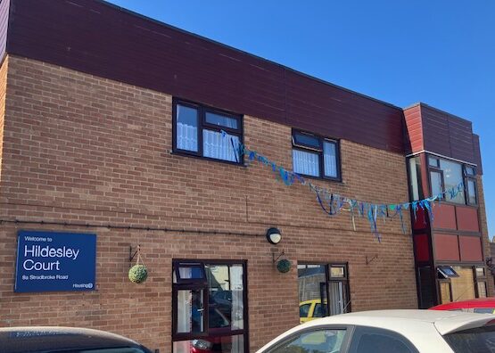 A length of blue bunting made from strips of different shades of blue fabric is suspended out of two second floor windows of a red brick building with the board saying Welcome to Hildesley Court