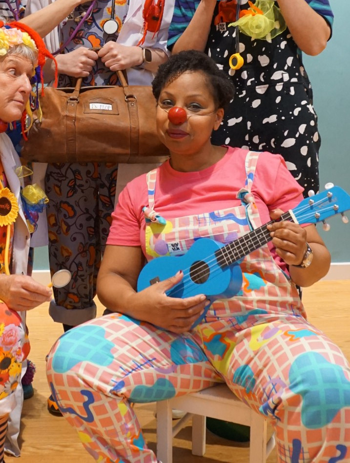 Clown Doctor Dr Kit Kat AKA actor, Katrina Beckford, wearing a red nose and playing the Ukulele.