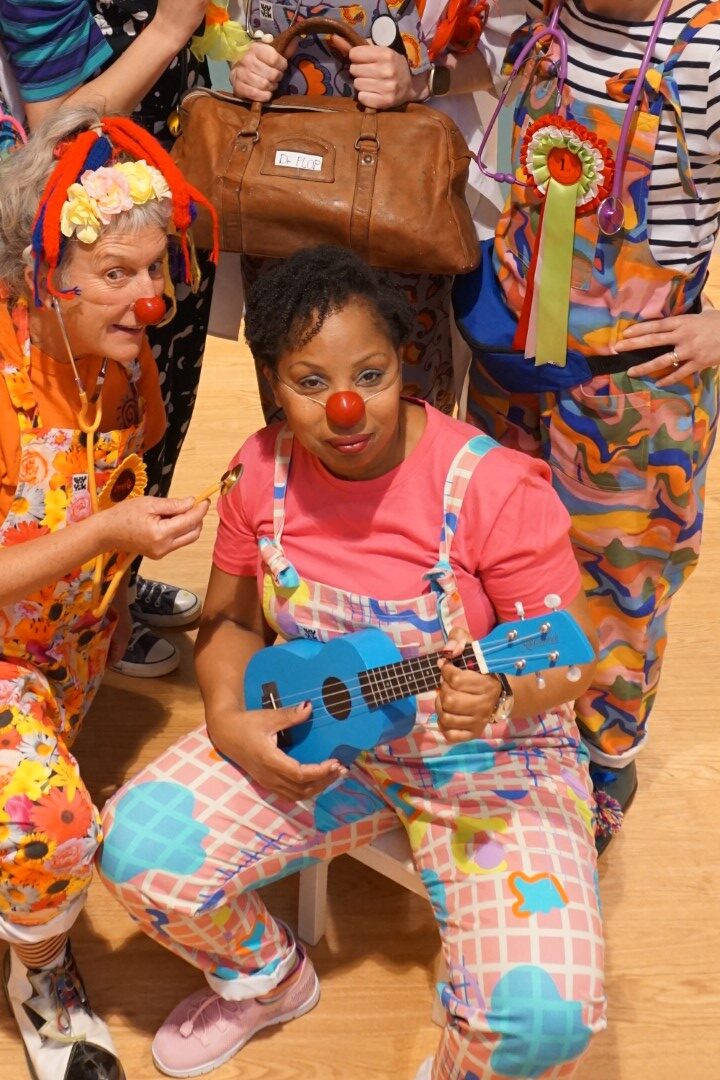 Clown Doctor Dr Kit Kat AKA actor, Katrina Beckford, wearing a red nose and playing the Ukulele.