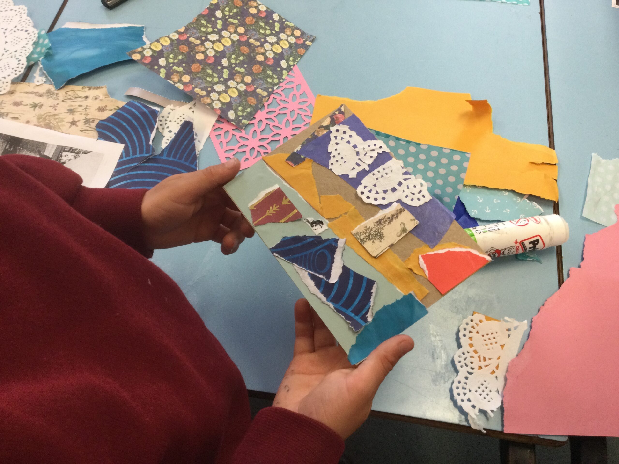 A child's hands hold a postcard, decorated with snippets of blue, yellow and pink paper and white doily. The child is obviously standing by a school table, on which are piles of decorative papers of all shapes and sizes and a glue stick