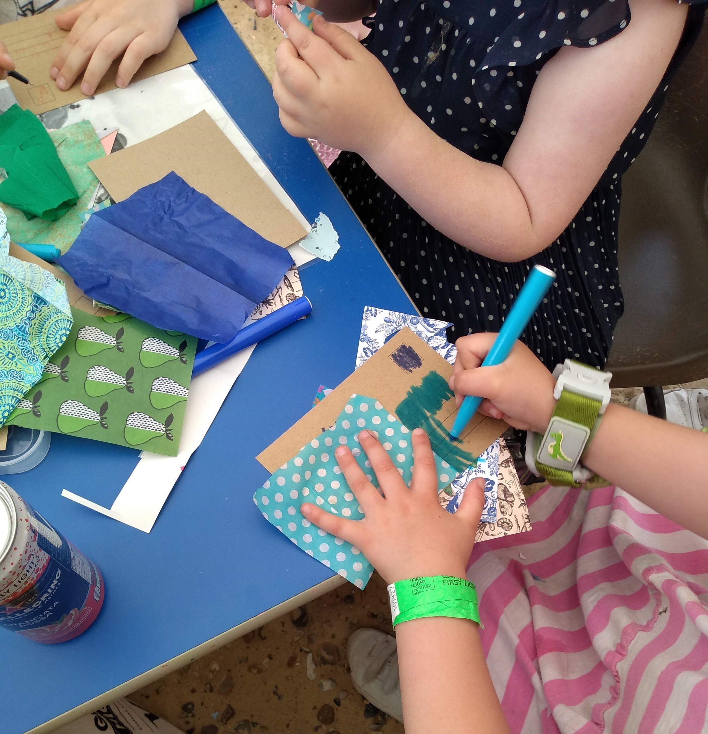 Children seated at a table. One is holding a piece of blue and white dotted tissue paper in place, whilst colouring beside it in blue felt tip pen. Another child is selecting from a pile of assorted paper