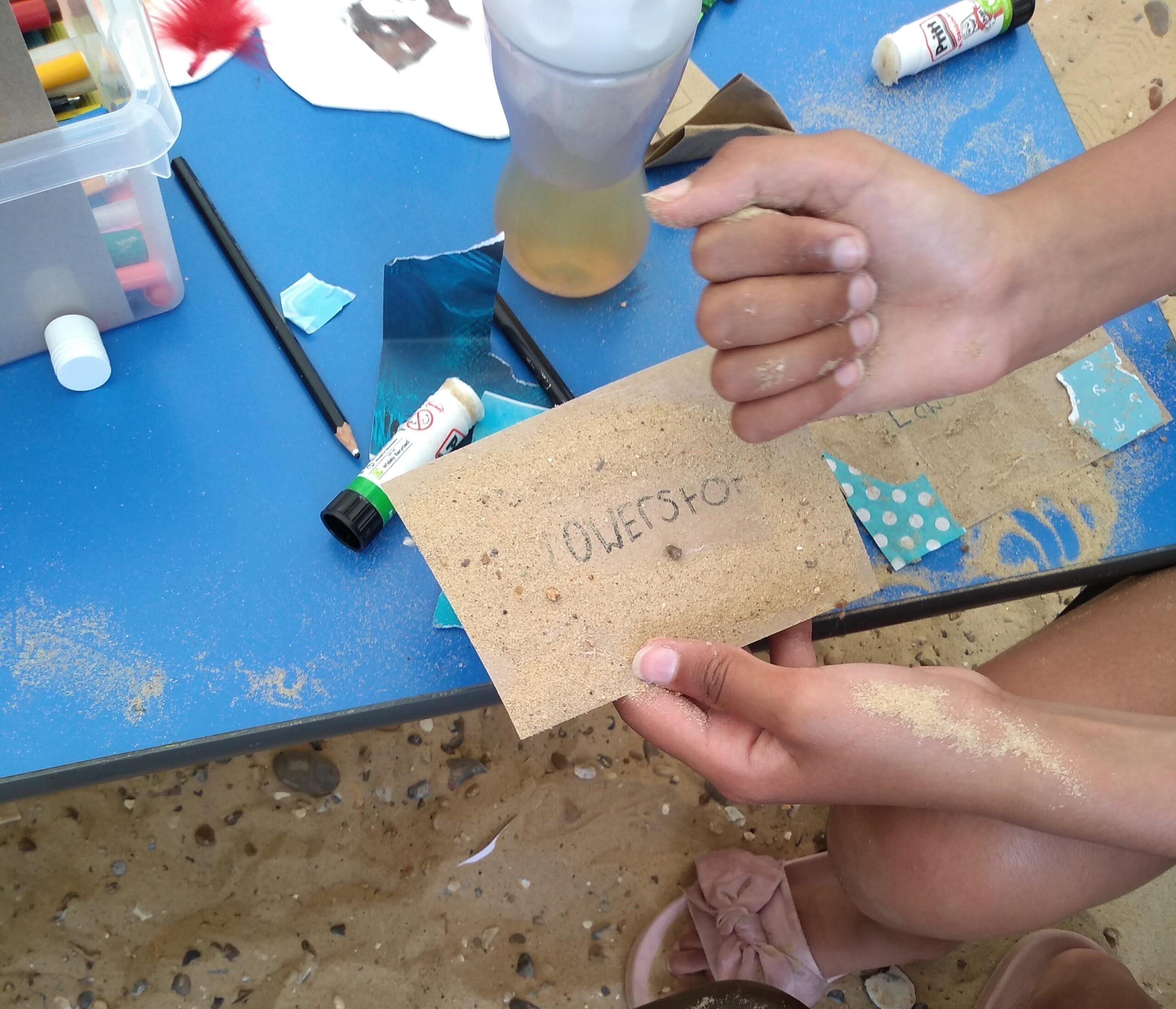 A pair of child's hands, holding a postcard and sprinkling beach sand onto the glued surface of a plain postcard with the word Lowestoft on it