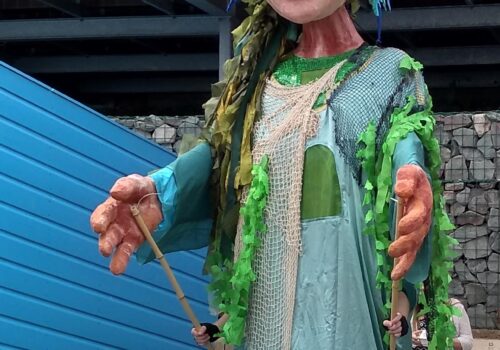 A huge puppet, powered by a person inside, using long poles to manoeuvre the large hands. The figure is dressed in long flowing net gown, drapes of green fabric resembling seaweed and has the rising sun as a crown