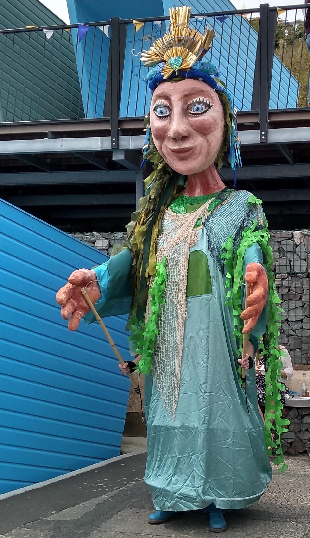 A huge puppet, powered by a person inside, using long poles to manoeuvre the large hands. The figure is dressed in long flowing net gown, drapes of green fabric resembling seaweed and has the rising sun as a crown