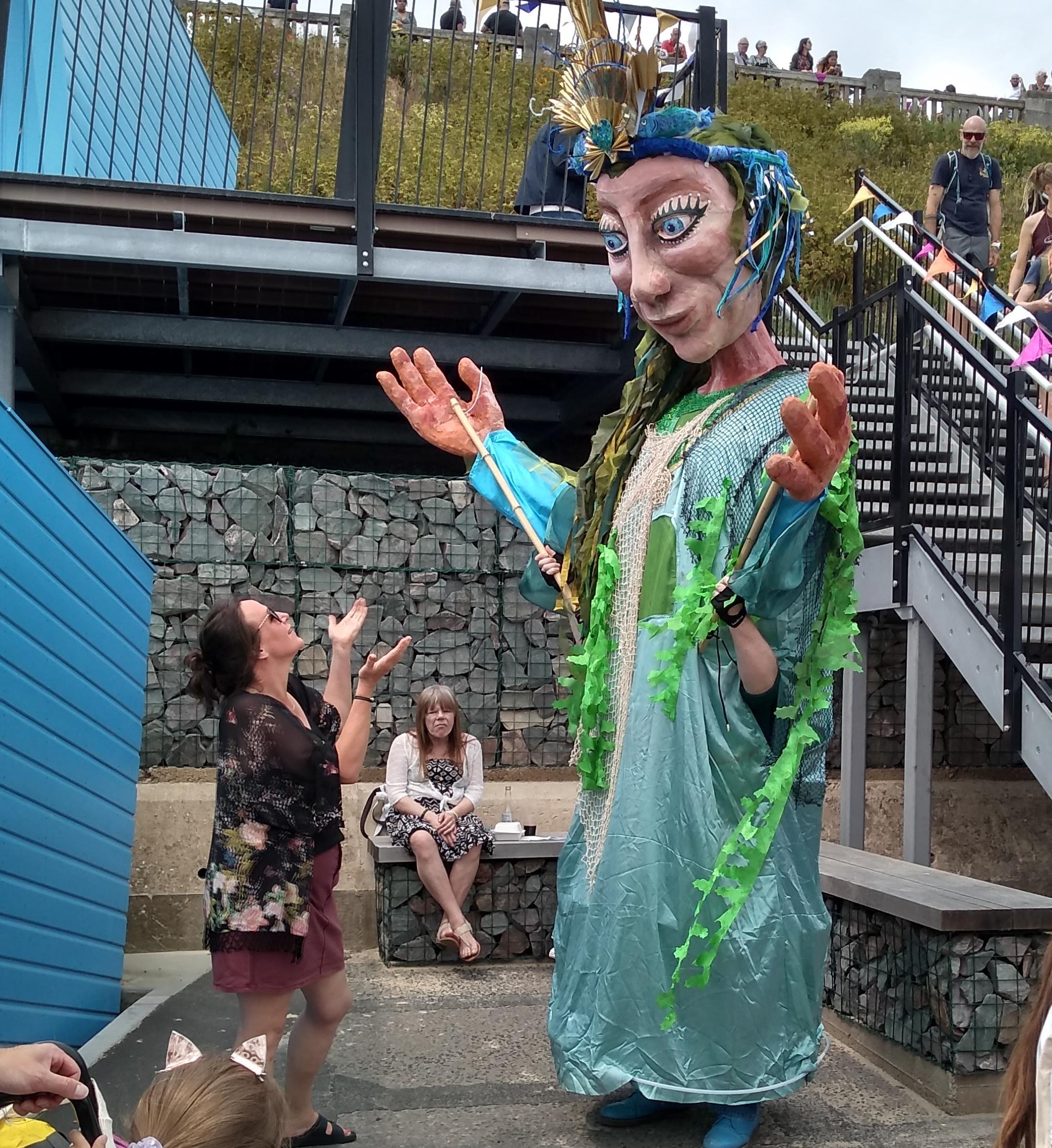 A massive puppet, twice human height, dressed in flowing aquamarine net gown and a rising sun crown holds out its hands, mirroring the actions of a member of the public standing in front of it.