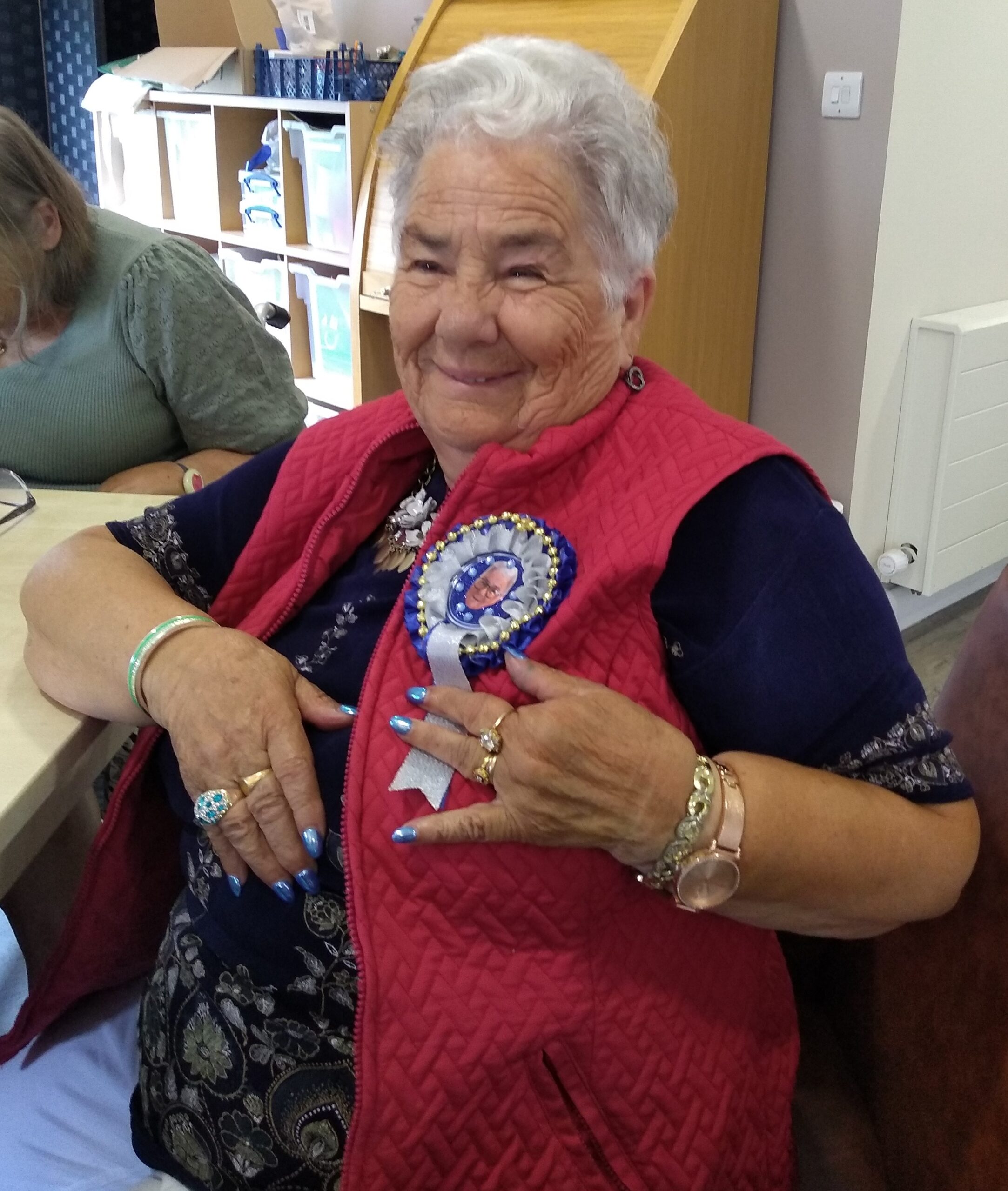 A person grins at the camera whilst proudly showing the rosette she's made, with a picture of herself and decorated with ribbons, beads and small blue gems