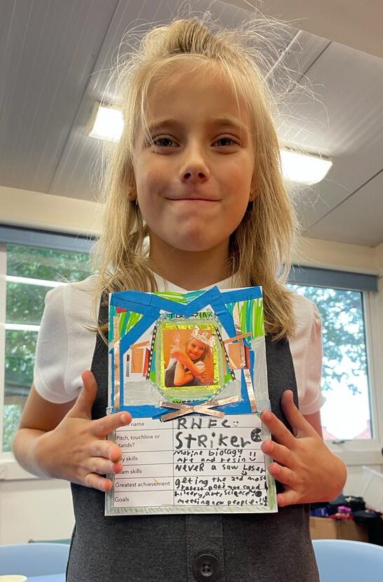 School child holding a decorated card on which she has listed her team skill as Never Being A Sore Loser