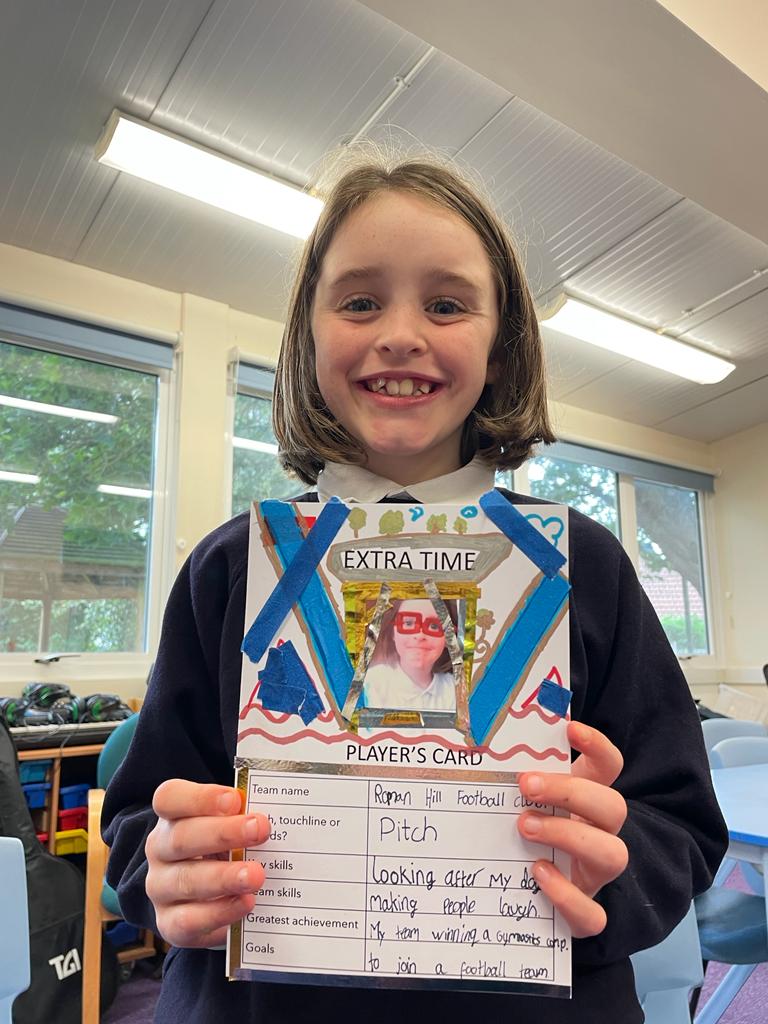 Child smiling at camera, holding her Extra Time Player's Card which she has decorated in blue and included a photo of herself