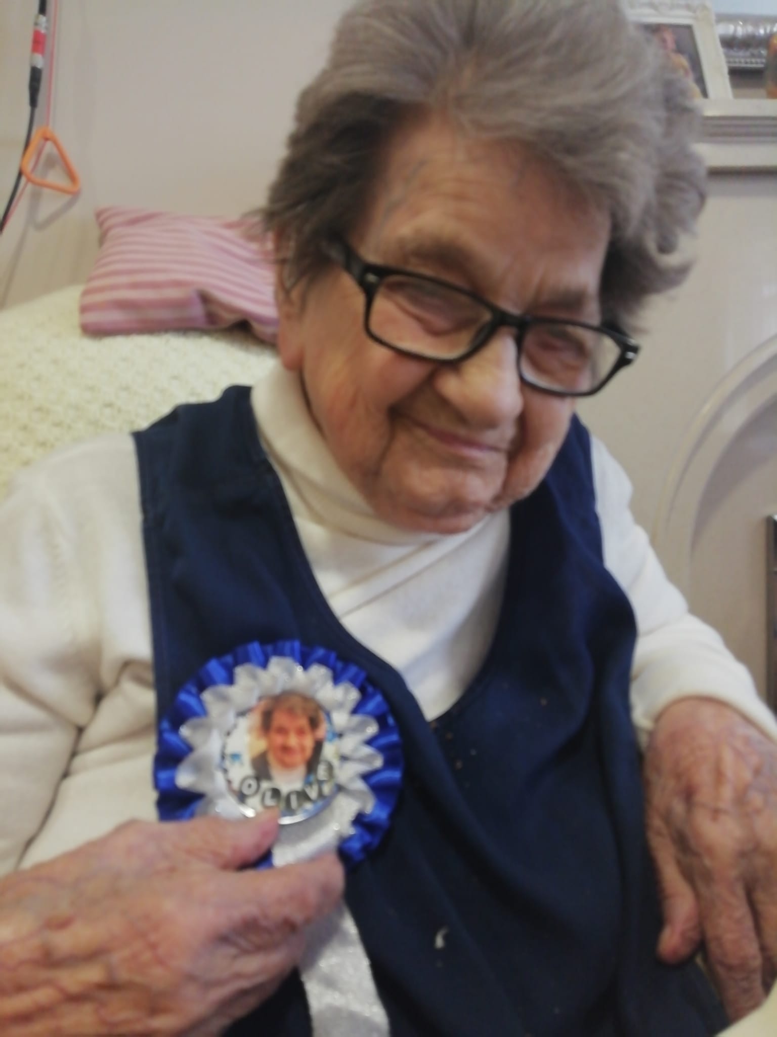 A lady in a white polo neck jumper and dark coloured waistcoat with a blue and white rosette pinned to it