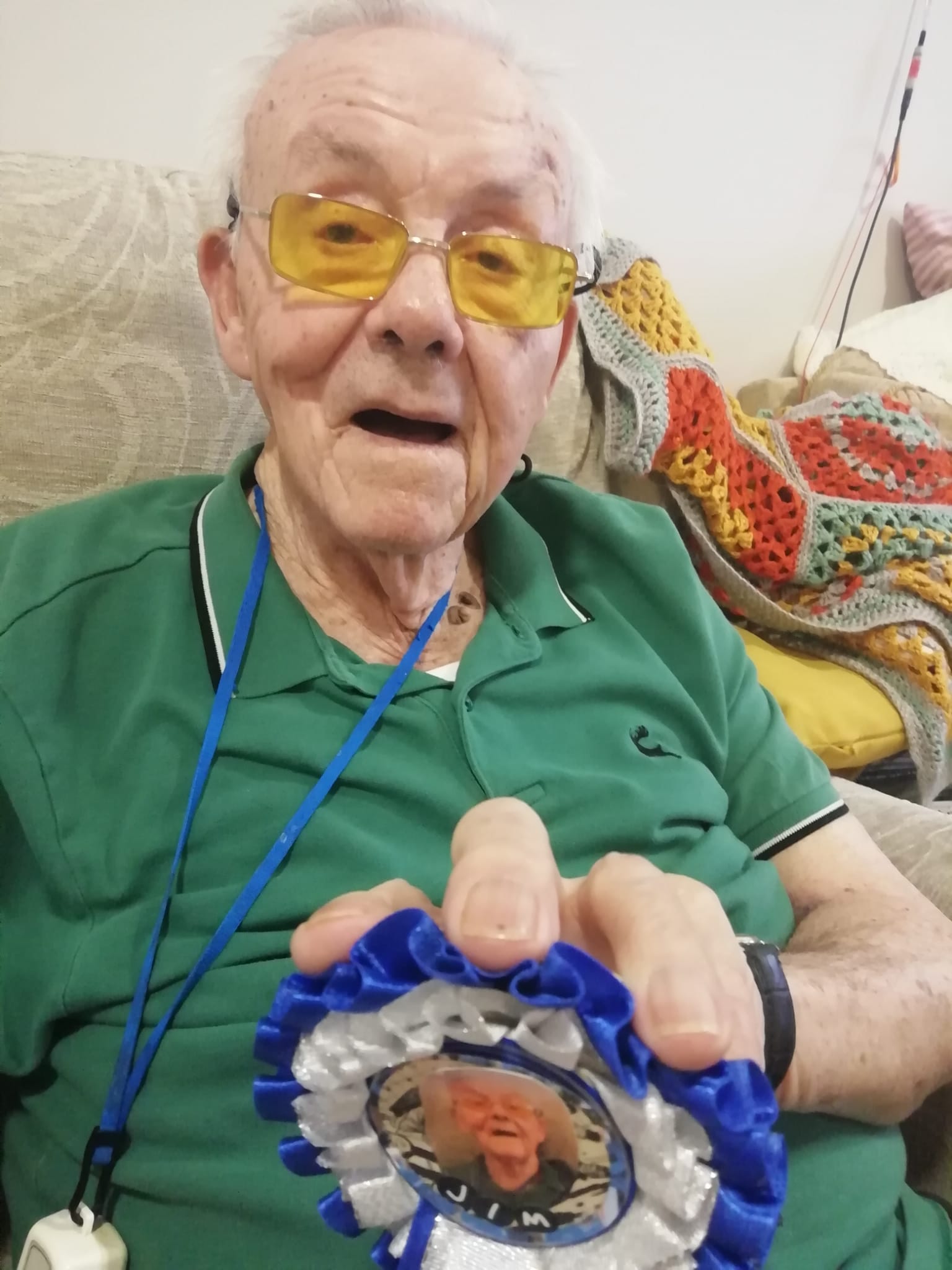 Gentleman seated, wearing a green short-sleeved top and holding a blue and white rosette he has decorated with an image and other details