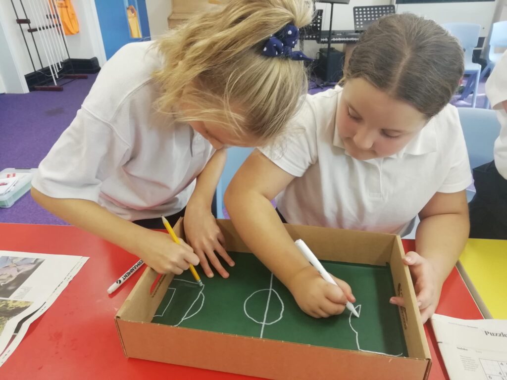 Two school children using pencils and a white marker to mark out a football pitch inside a cardboard box