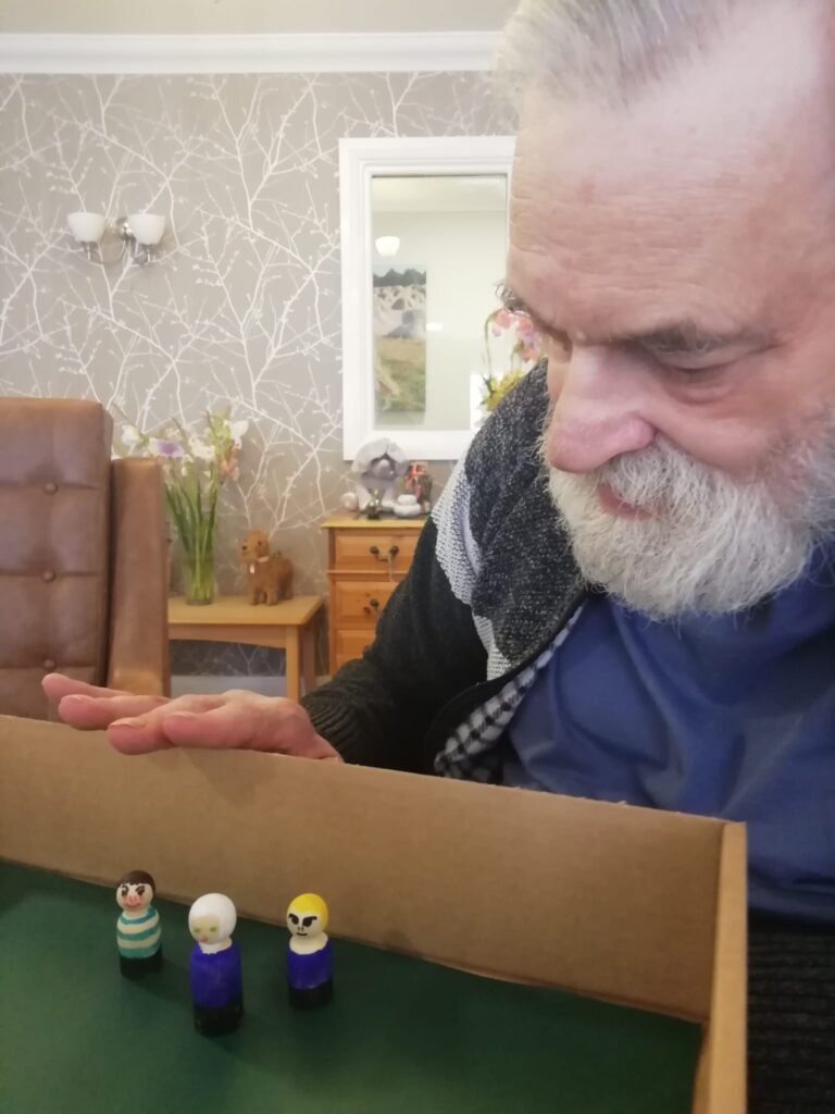 A gentleman looking closely at three small wooden figures inside a cardboard box fashioned to look like a football pitch