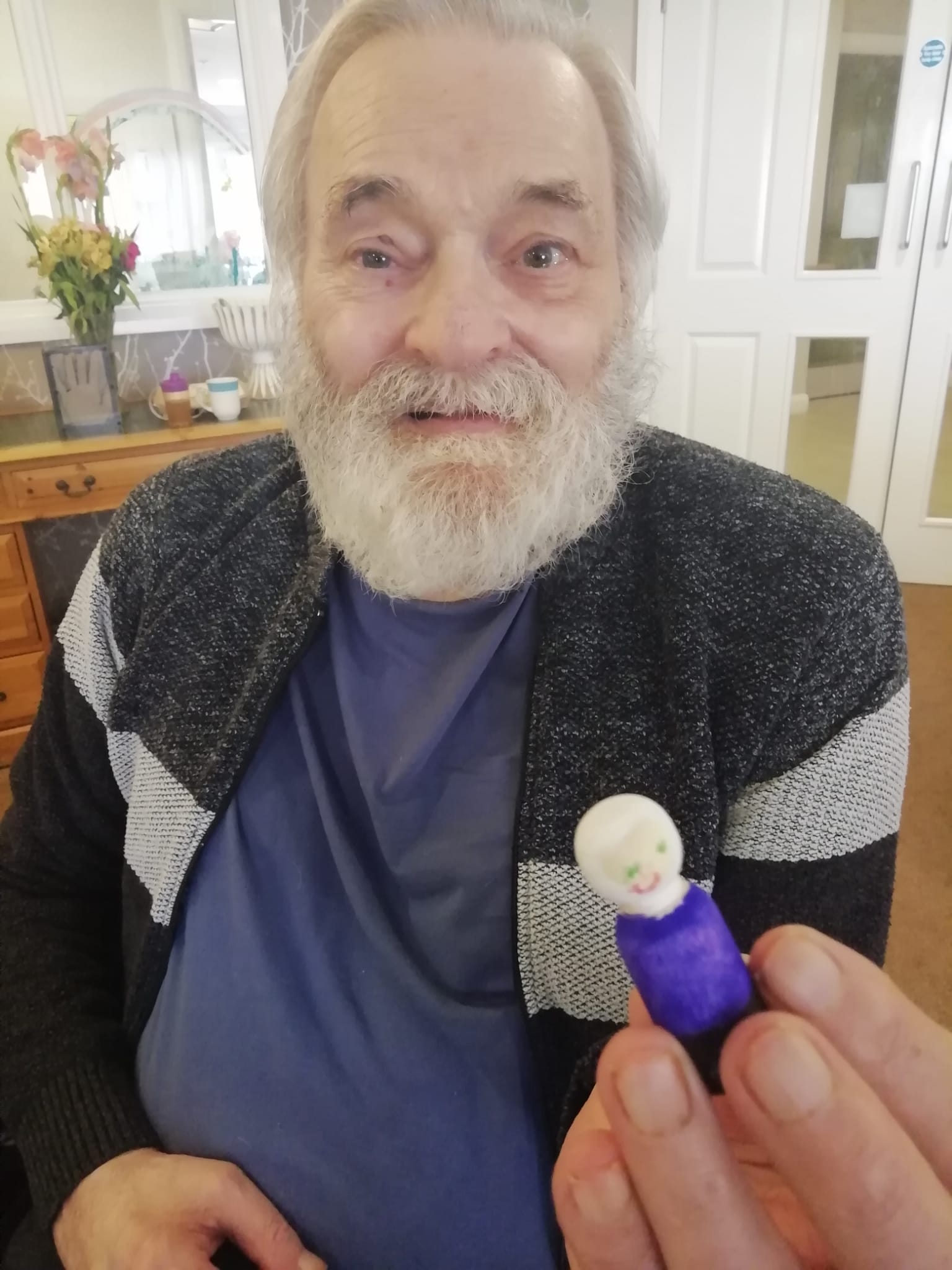 A gentleman in a blue T-shirt and grey cardigan holds a small, hand decorated wooden figure up to the camera