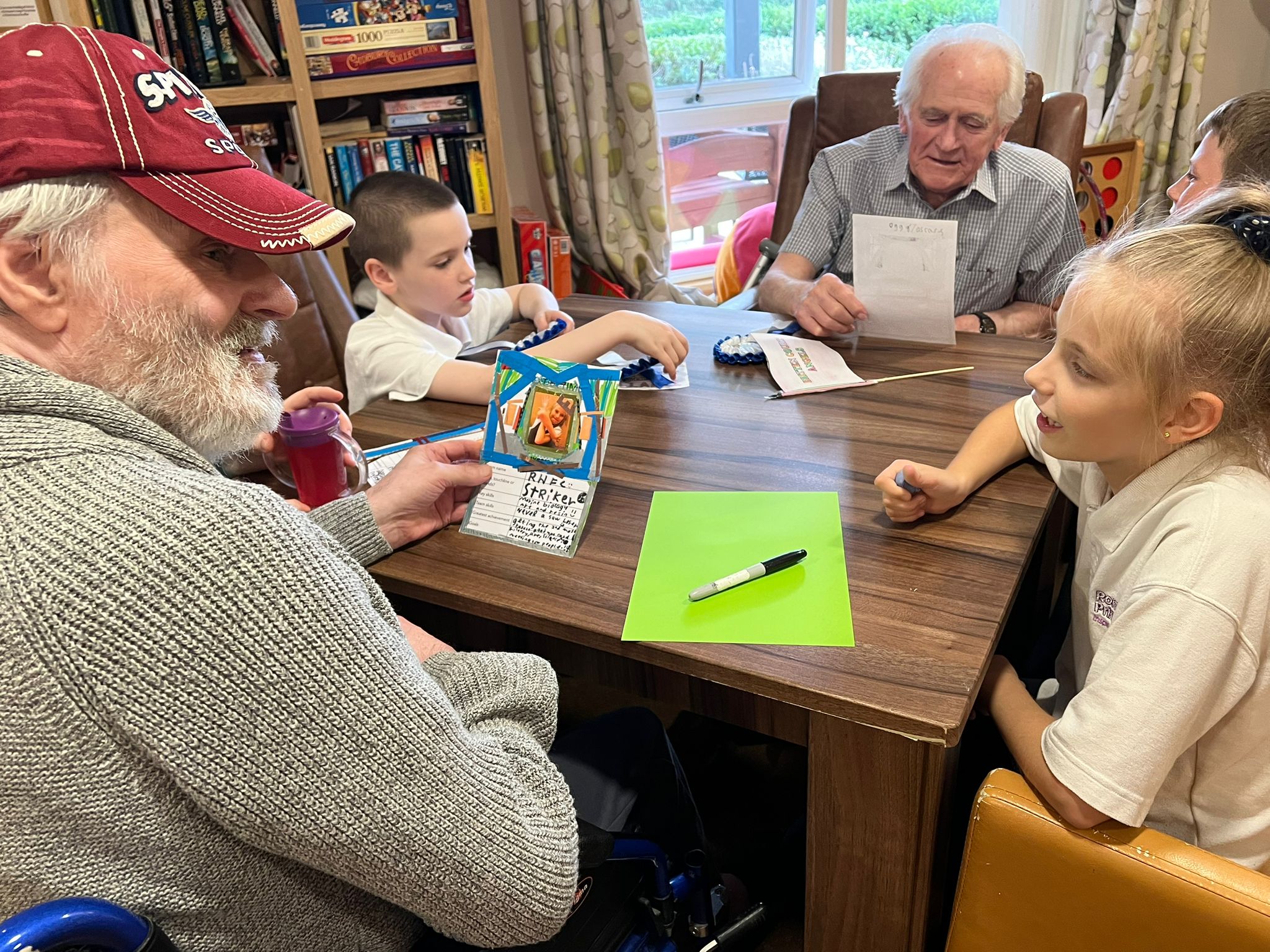 Two gentlemen, one wearing a baseball cap, sit at a table looking at some decorated cards made by the children seated with them