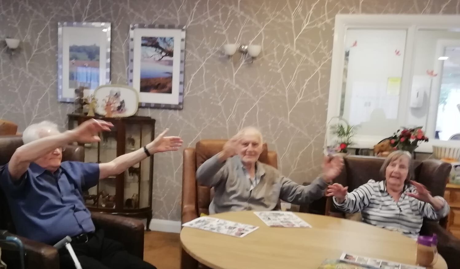 Three adult, seated in easy chairs around a wooden table, waving their arms in the air