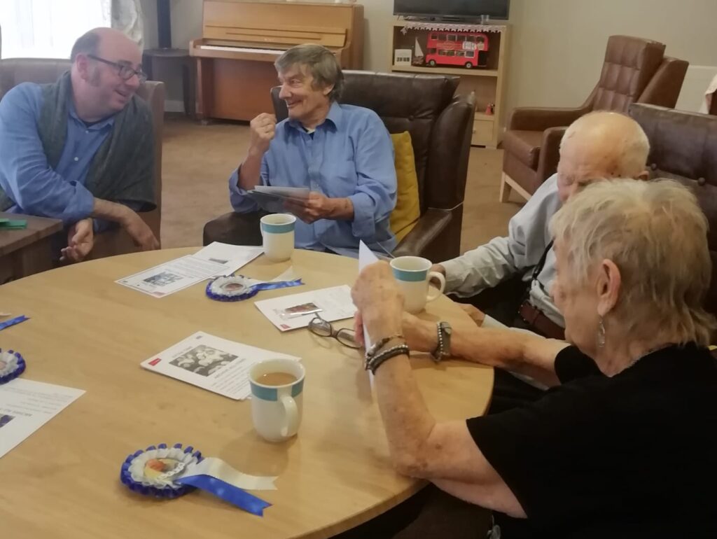 Four people seated around a table, drinking mugs of tea and having a chat. On the table are blue and white rosettes and information cards