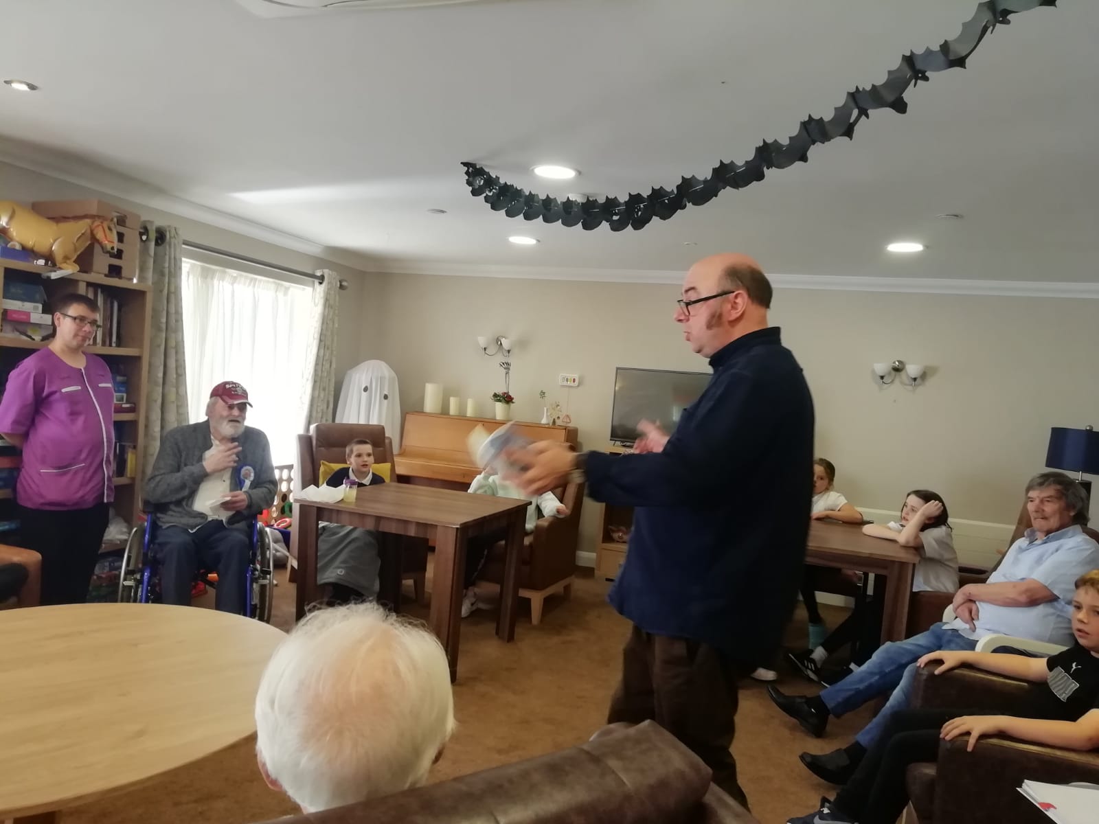 A large room in which a man stands surrounded by adults and children seated in a circle around him as he reads from a book. Suspended from the ceiling is a halloween decoration of a length of black paper bats