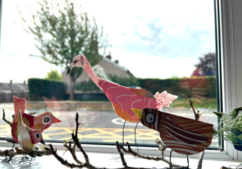 Artworks of colourful birds perched on a branch, displayed in a window.