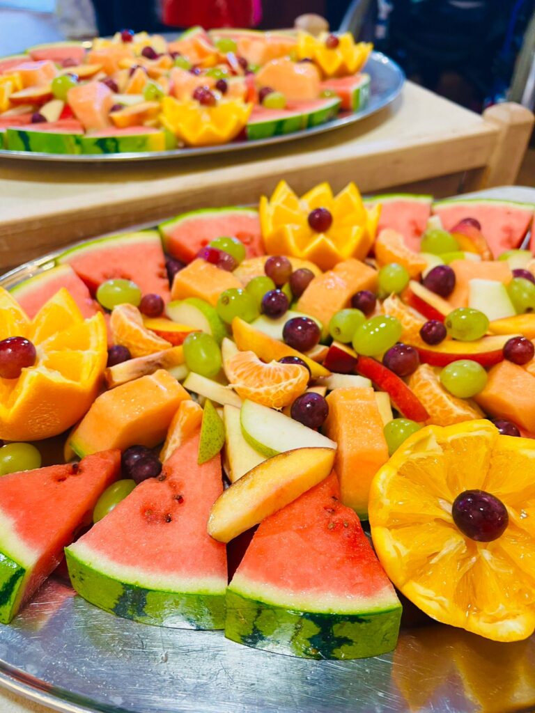 Two silver-coloured platters with a mixture of sliced melon, orange, apples and green and black grapes