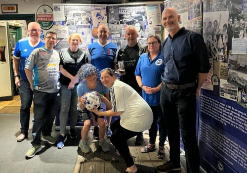 A group of adults, some wearing Ipswich Town Football Club T-shirts standing in front of displays of Town memorabilia. In the foreground, two women, one in a blue and white glittery wig, crouch holding a football