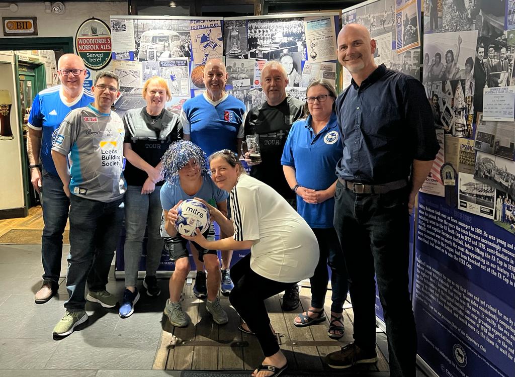 A group of adults, some wearing Ipswich Town Football Club T-shirts standing in front of displays of Town memorabilia. In the foreground, two women, one in a blue and white glittery wig, crouch holding a football