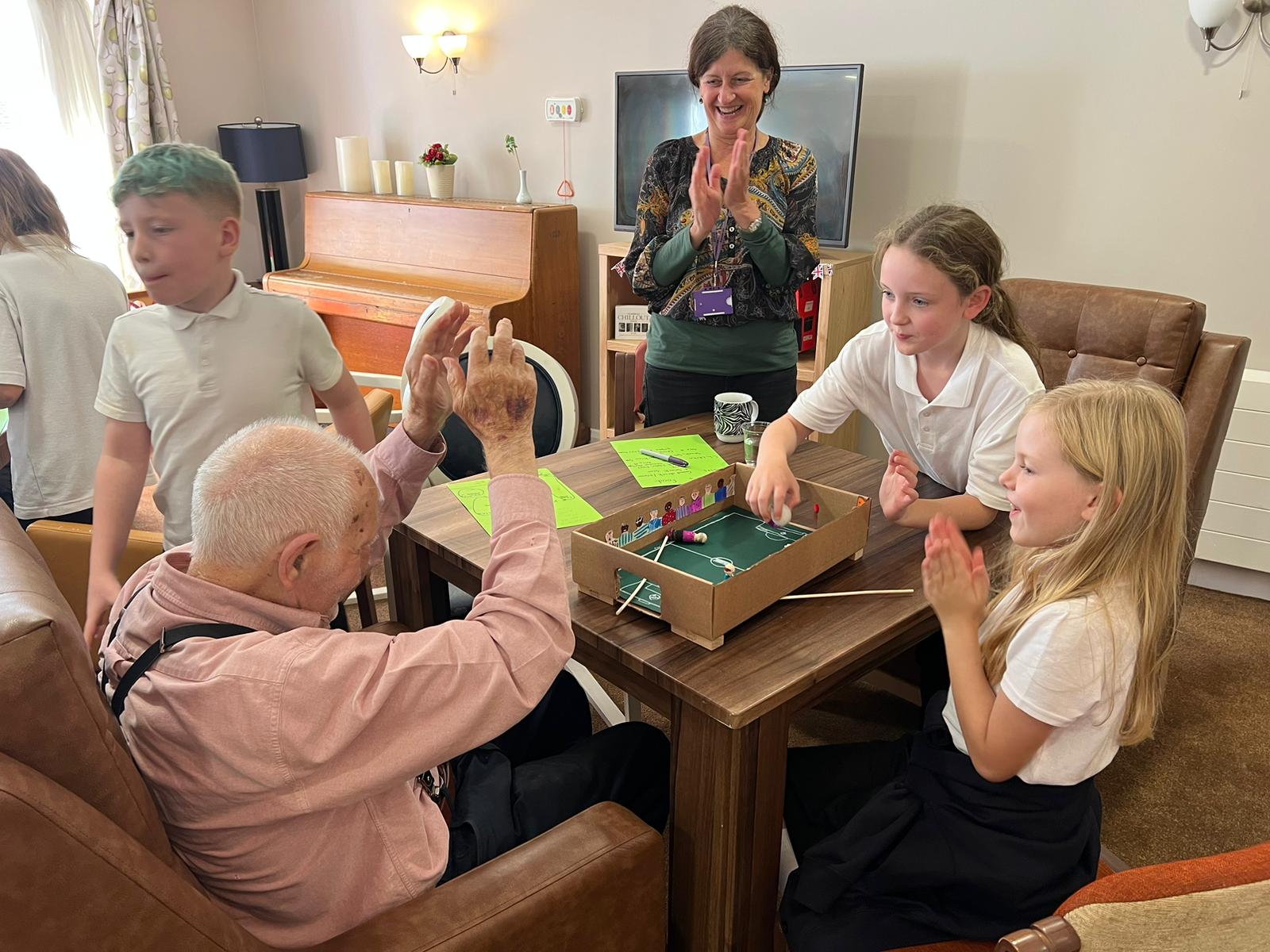 A gentleman seated, arms raised in the arm obviously in delight. On a table in front of him is a magnetic football game and three school children surround him, clearly taking part in the game. A member of staff watches on, applauding