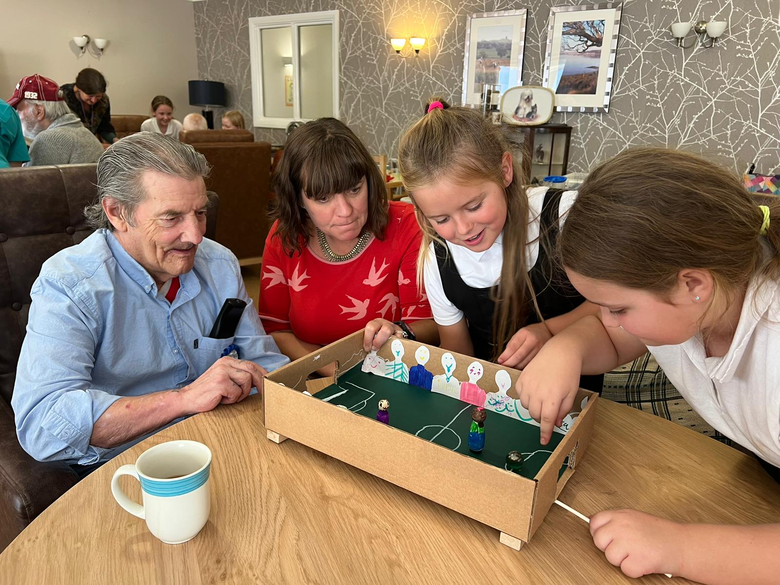 Two adults watch keenly, as two school girls adjust a small magnetic figure inside a cardboard football pitch