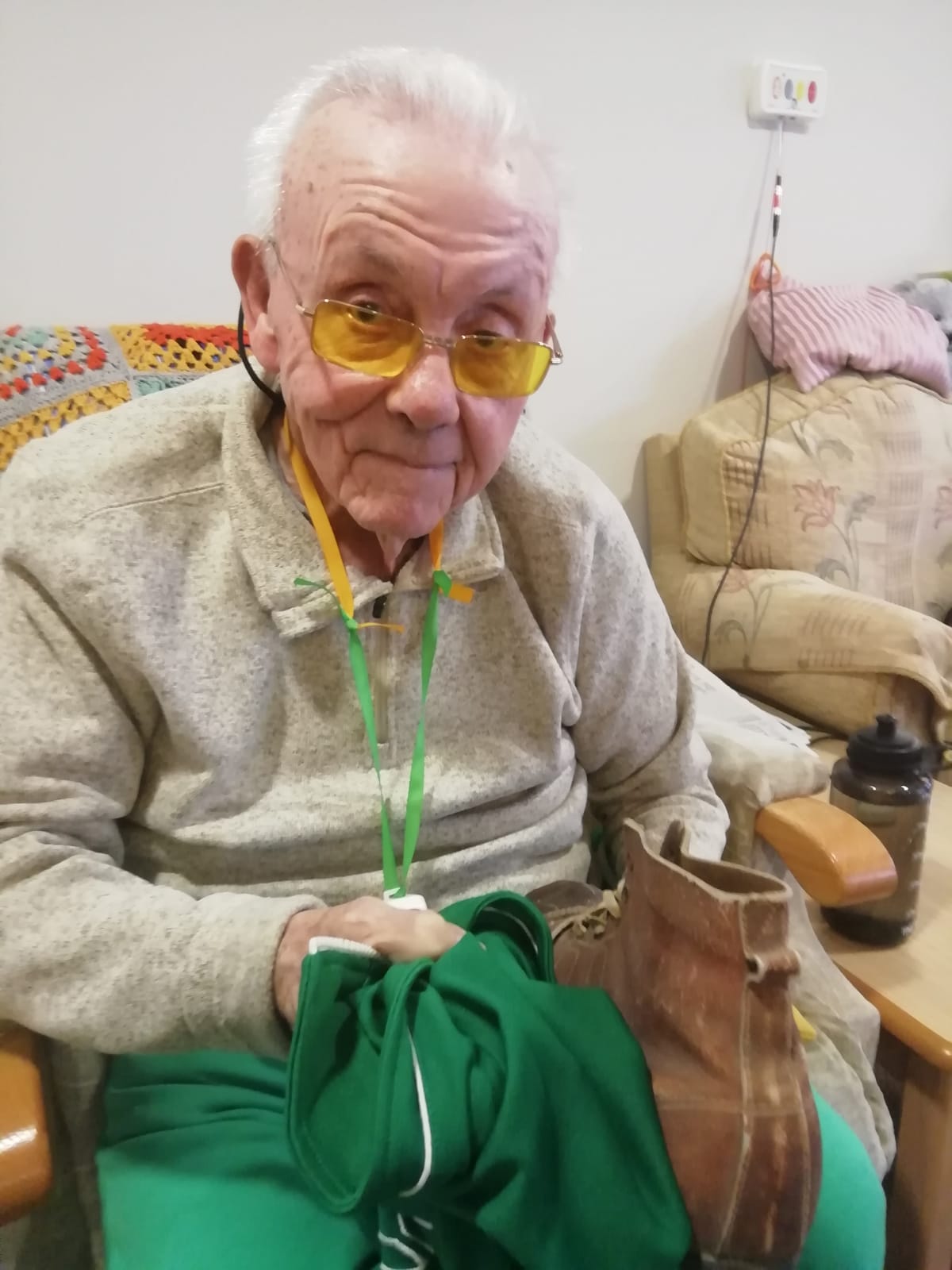 A gentleman seated in an armchair, holding an old leather football boot and a green football top