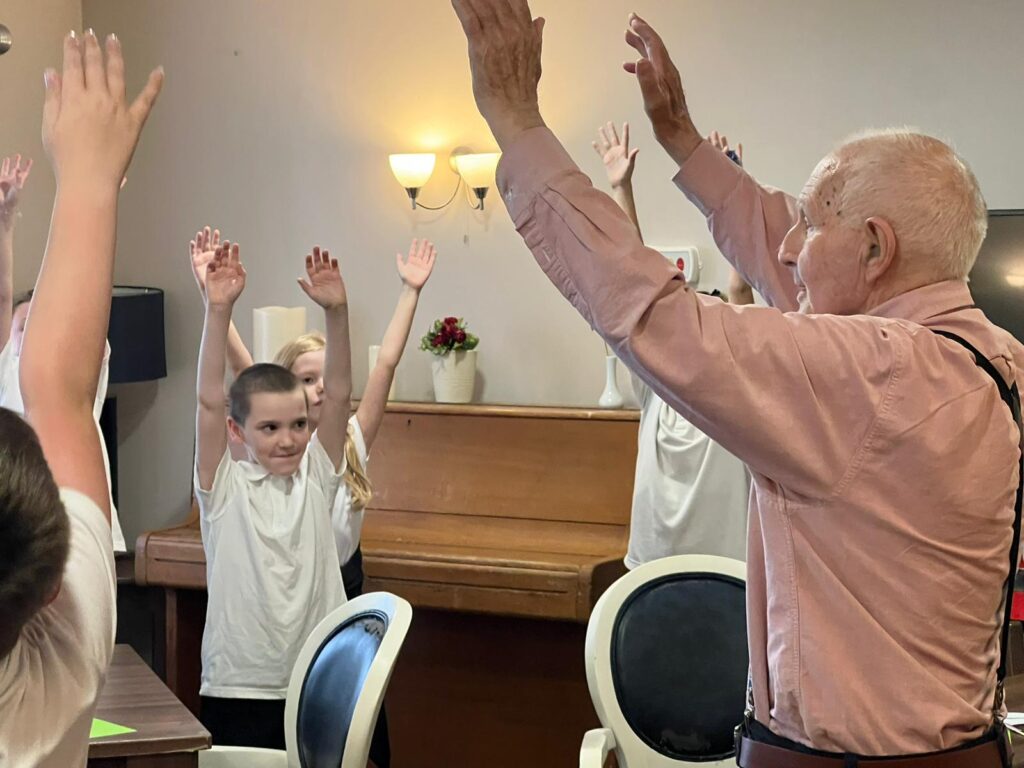 A gentleman stands, arms raised in the air, as a group of school children copy his movements
