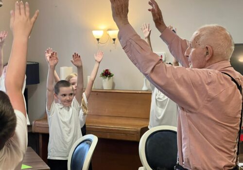 A gentleman stands, arms raised in the air, as a group of school children copy his movements