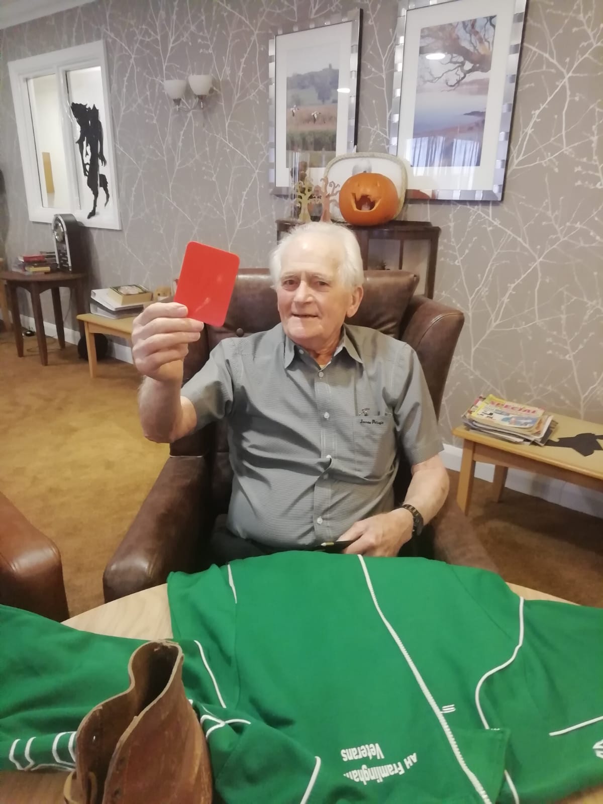 A gentleman seated at a table on which a green football top is spread out beside a leather boot. The man is holding up a red card