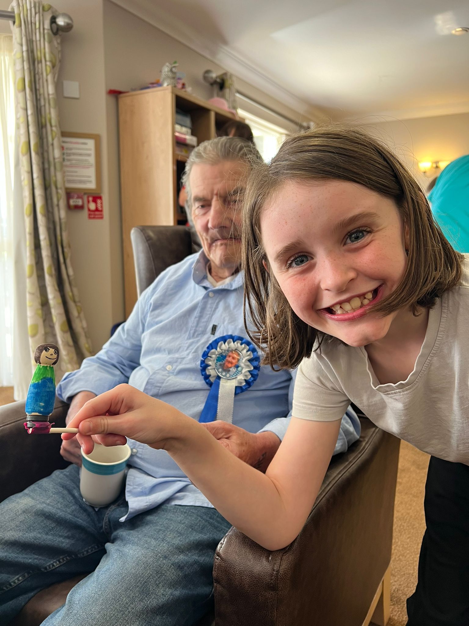 A child smiles broadly at the camera, holding a small football character on a stick. Behind her, seated in a chair, is a gentleman wearing a hand-decorated blue and white football rosette