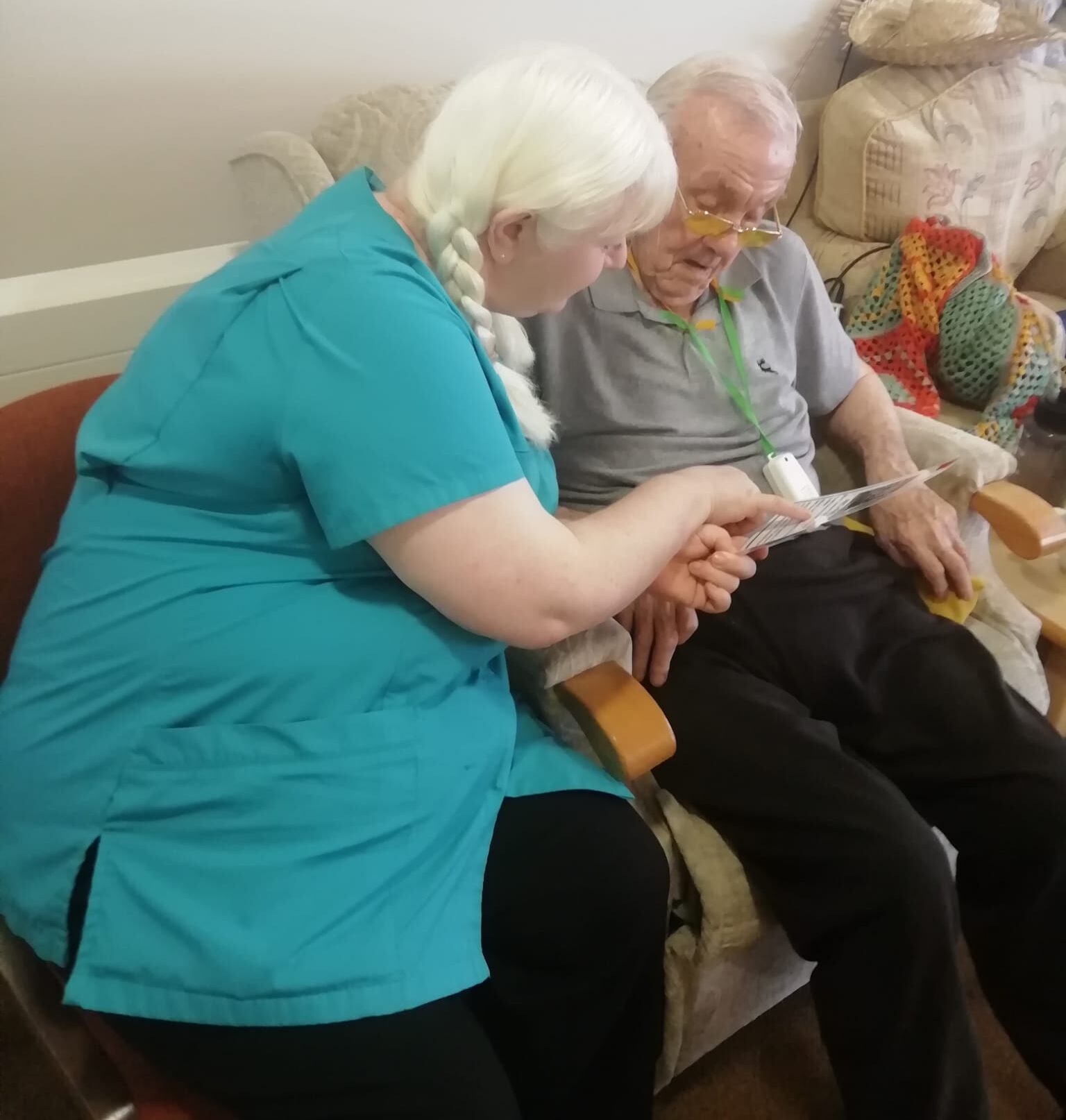 A woman in a turquoise uniform top and blond hair in long braided pigtails is showing a gentleman a picture of something and they are discussing it together