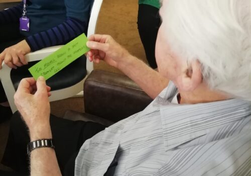 A back view of a gentleman seated in an armchair, holding a strip of green paper with words written on it