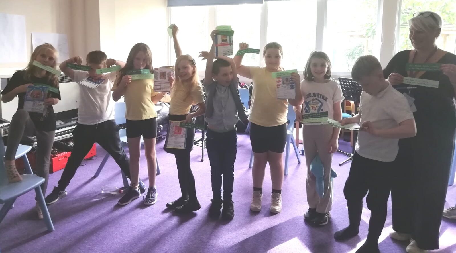 A group of school children holding up cards and lines of text to camera, with member of staff looks on