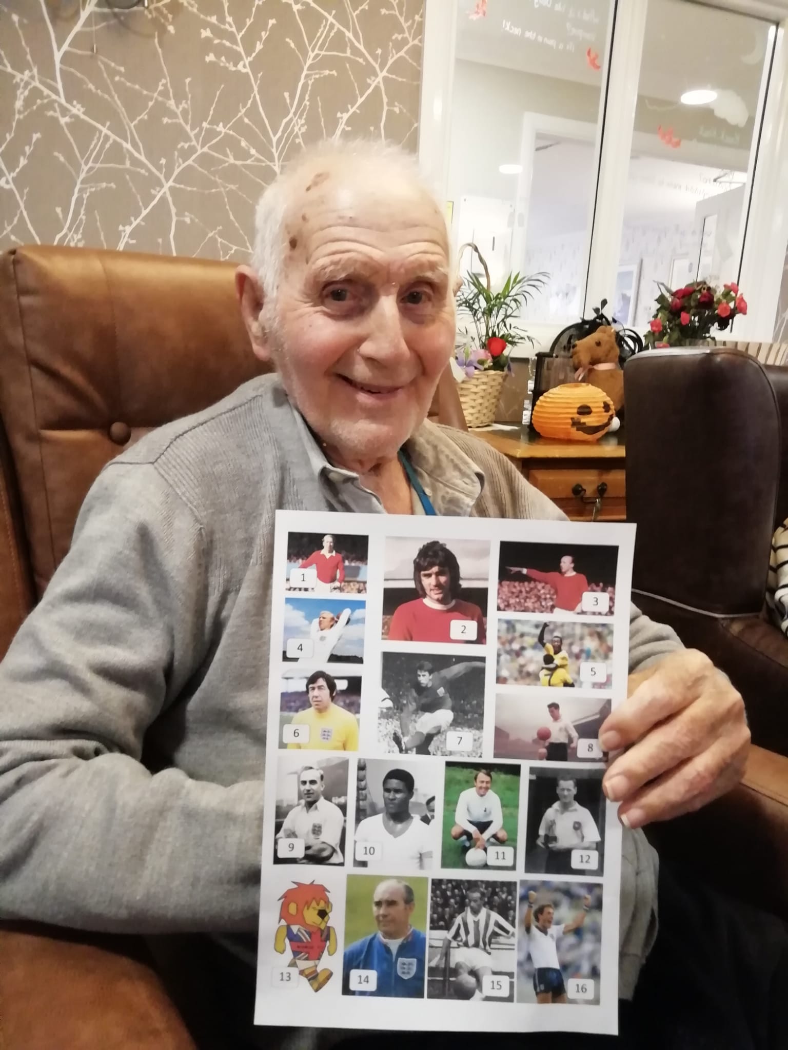 A gentleman smiles directly to camera. He's seated and holding a football quiz sheet featuring pictures of many famous footballers