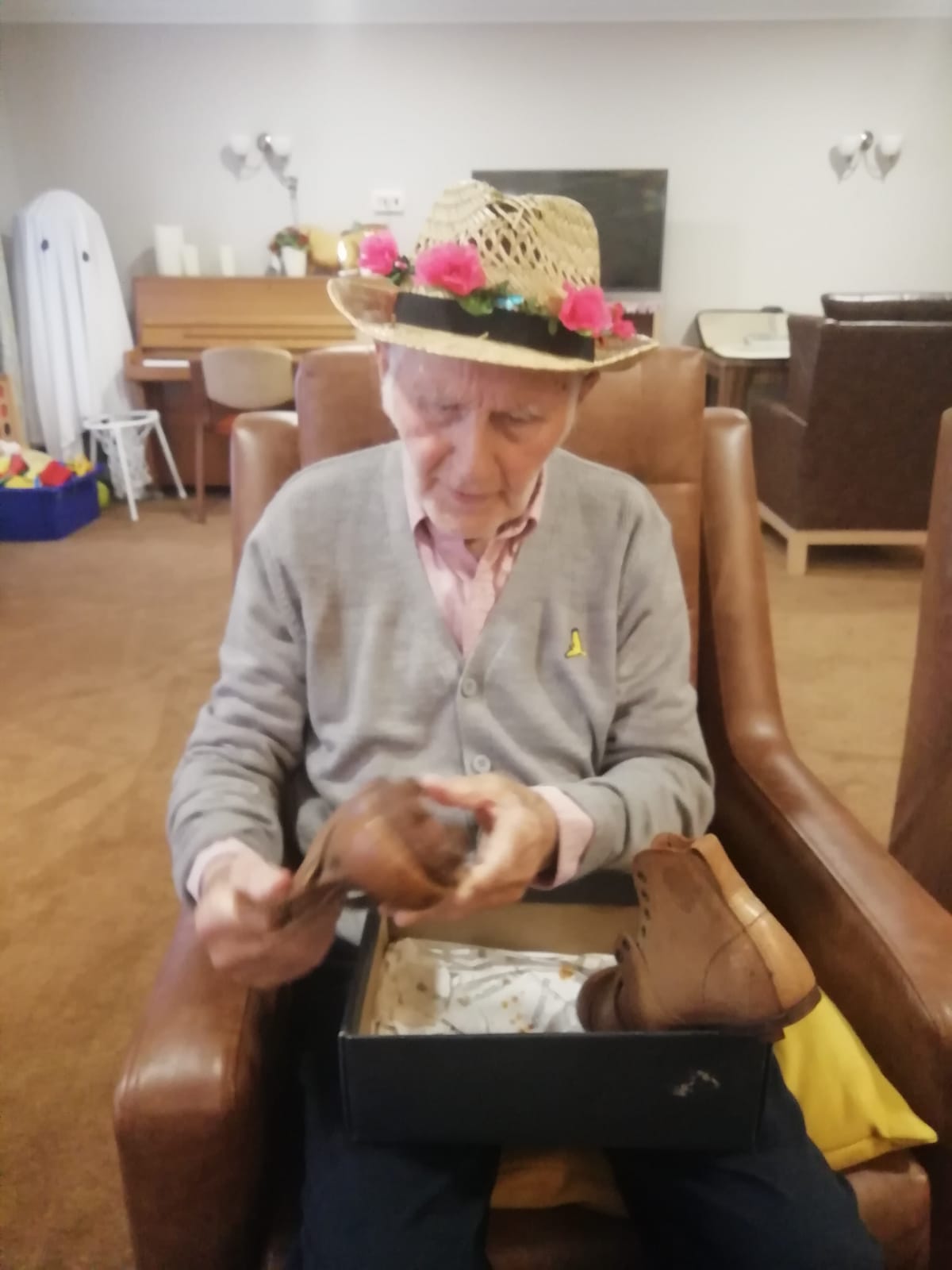 A gentleman seated in a brown armchair and inspecting a pair of leather football boots. He is wearing a straw hat with pink flowers around the brim